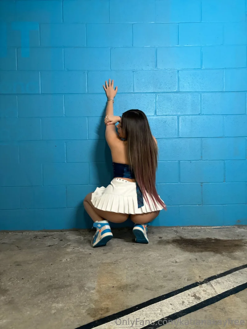Hot Katiana Kay squatting against blue wall in cheerleader skirt and crop top