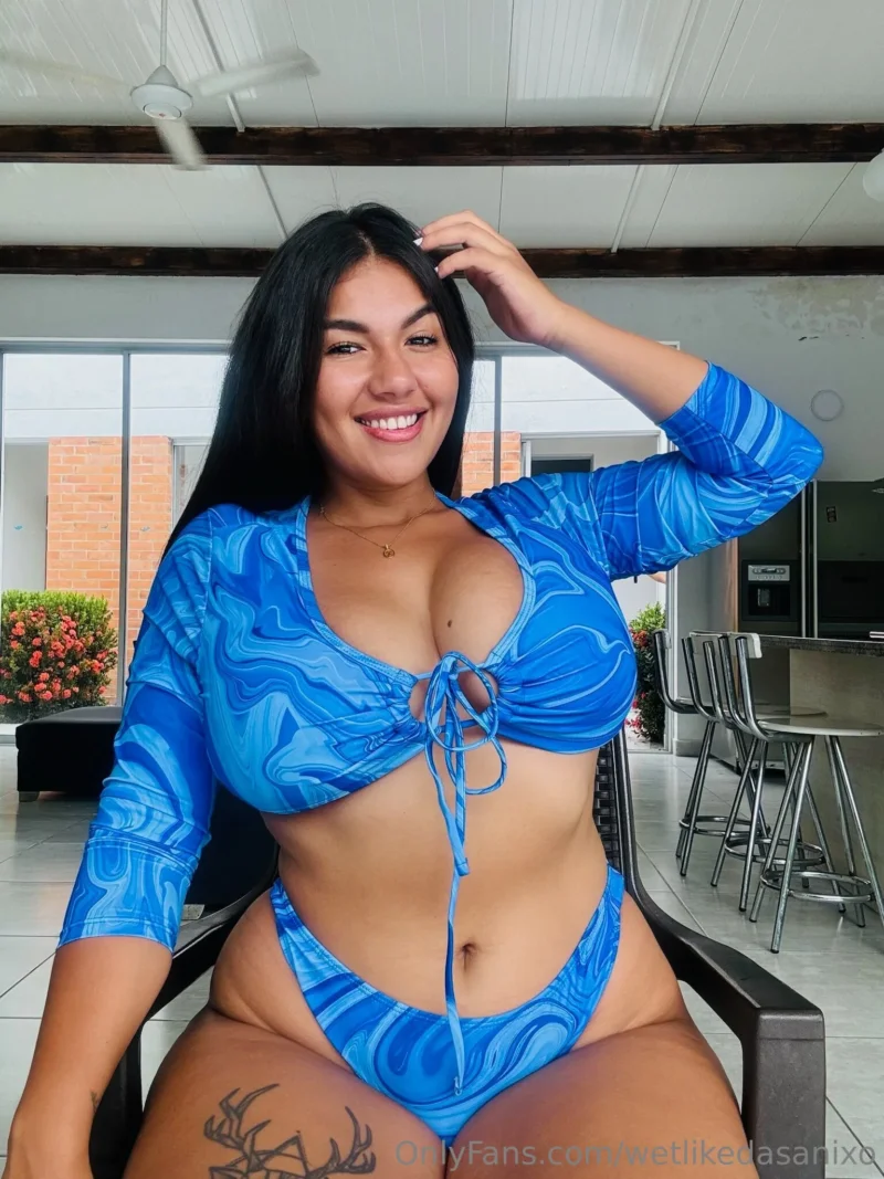 Seductive Dasani Delgado in blue swirl bikini top, smiling with hand in hair, tattoo on thigh