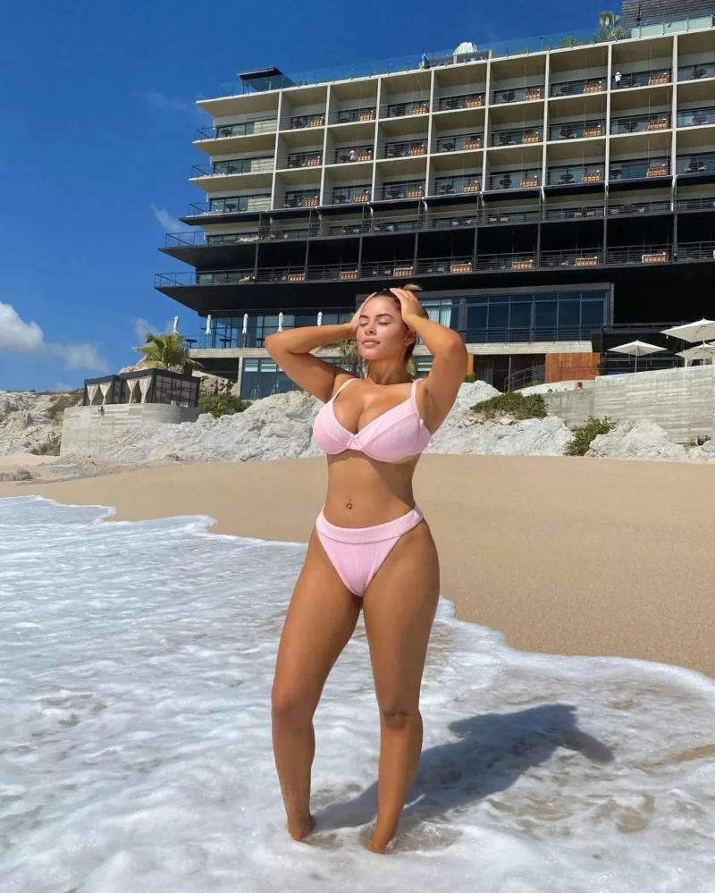 Busty Noelia Ramirez in pink bikini standing in ocean waves with hands on head