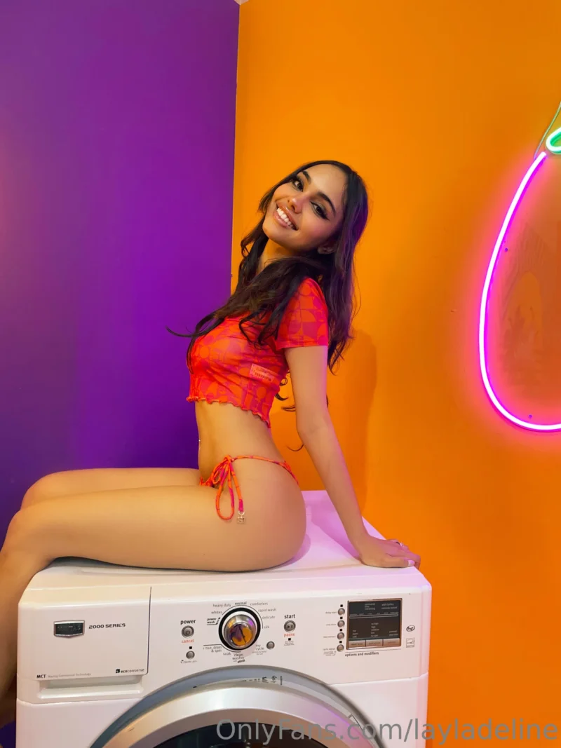 Seductive Layla Deline straddling a washing machine in orange bikini, smiling at camera