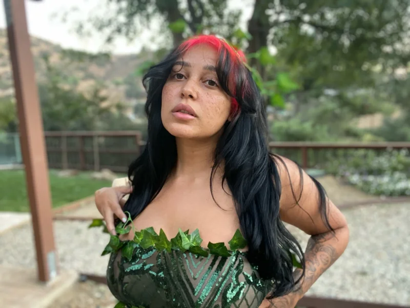 Curvy Emily Cheree in leafy green top, posing outdoors with one hand in her hair