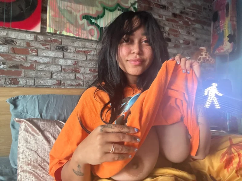 Seductive Emily Cheree lifts her orange tee to expose her tits on bed