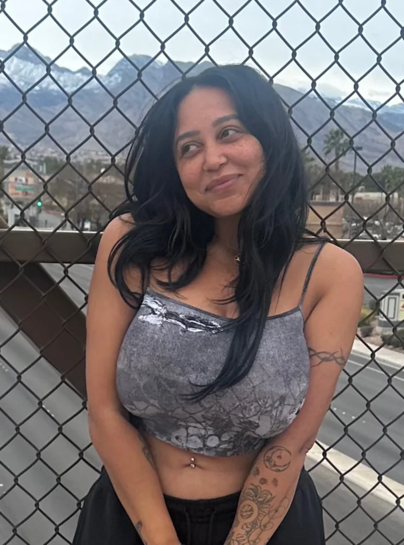 Sultry Emily Cheree leaning against chain link fence in grey crop top, showing off her belly button
