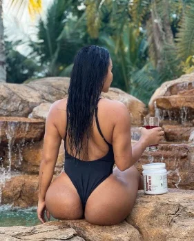 Seductive Dolly Castro in black one-piece bikini sits on rocks by waterfall holding drink