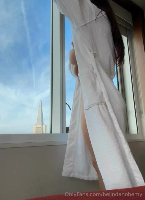 Busty Belinda Nohemy in white robe lifts leg at window showing thigh and ass