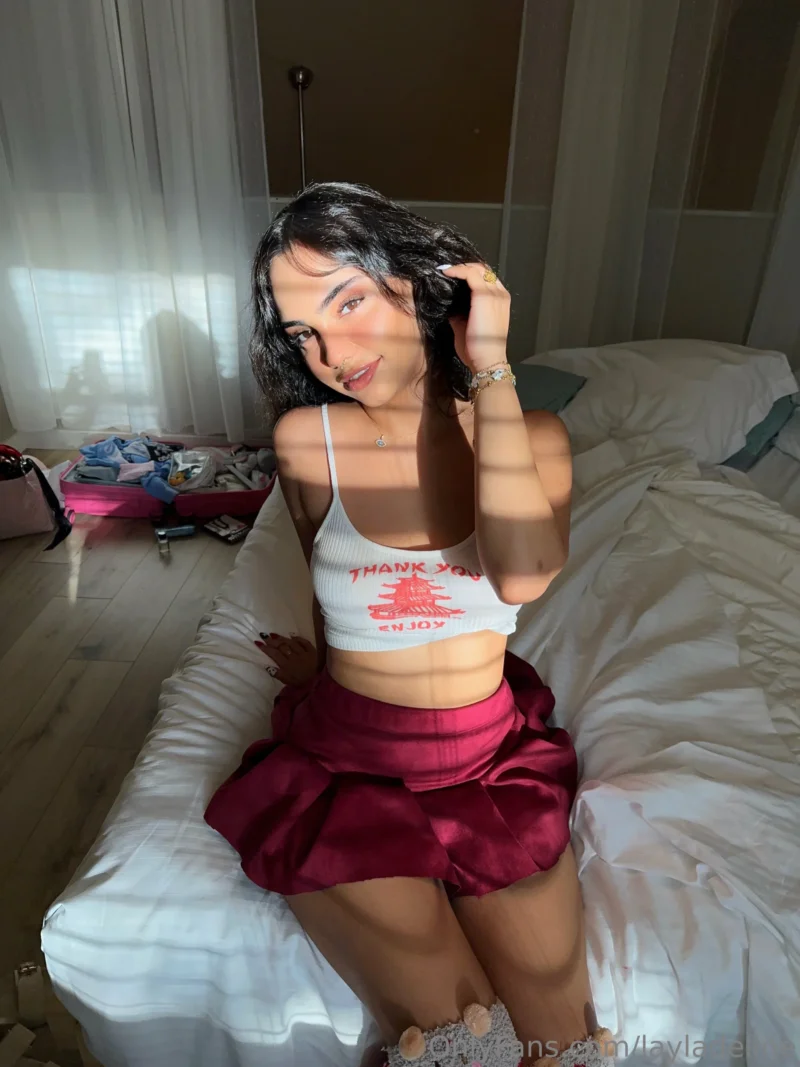 Seductive Layla Deline lounging on bed in white crop top and red skirt, hand in hair