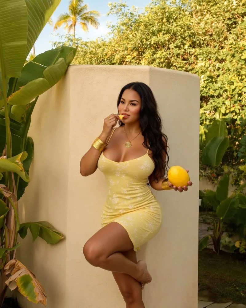Curvy Dolly Castro in a yellow dress eating fruit by a wall with palm trees
